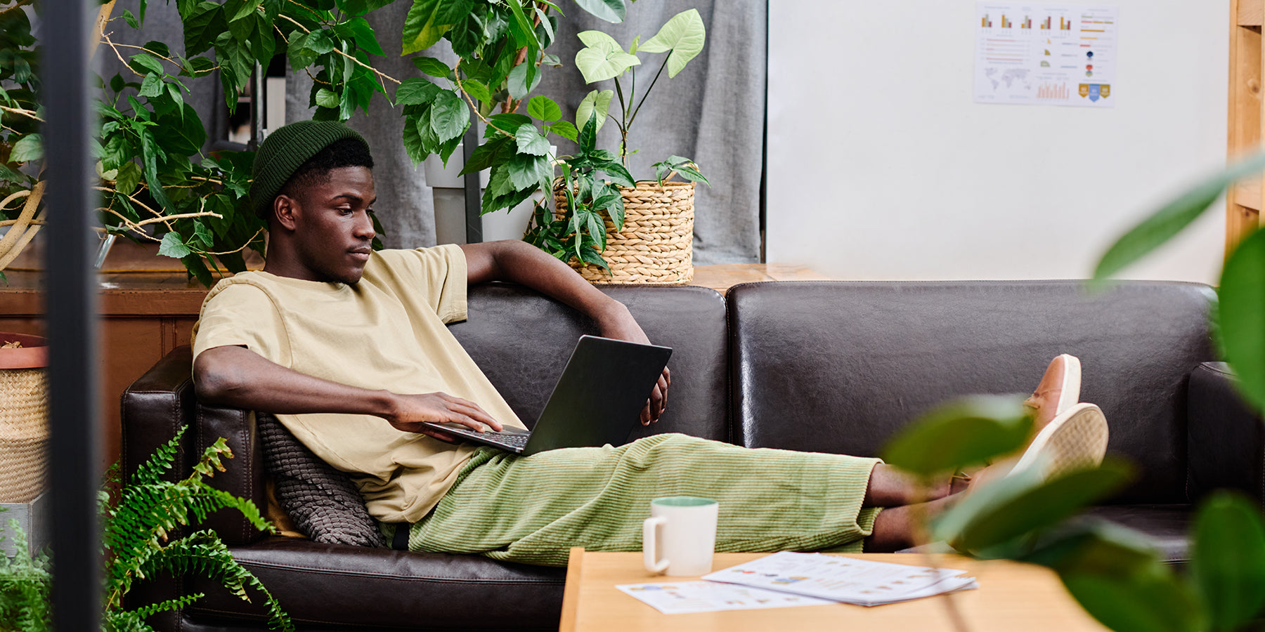 A man sitting on a couch with a laptop in an office surrounded by plants.